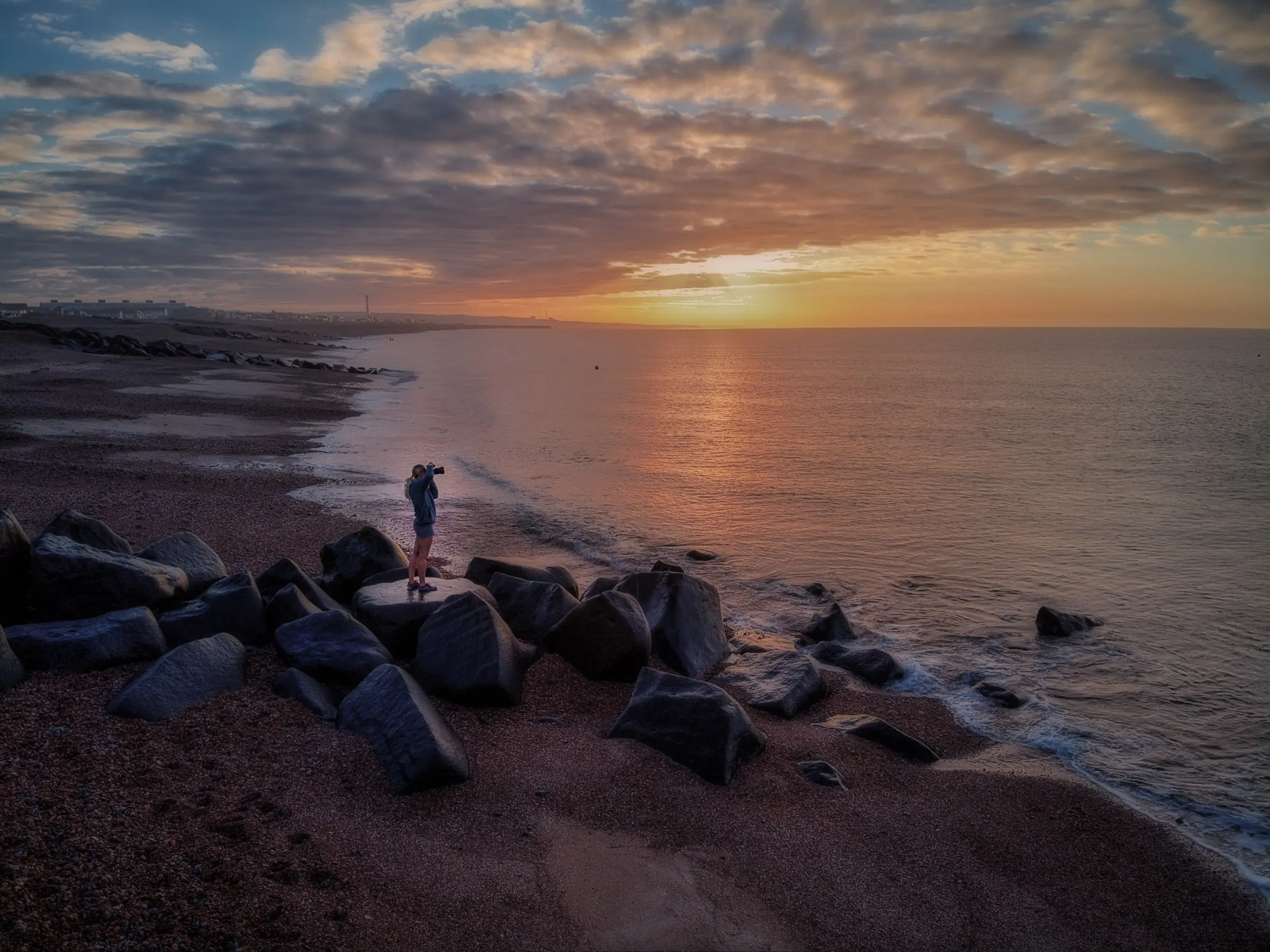 Petra stands on the rocks on Shoreham beach at sunrise, photographing the sea.