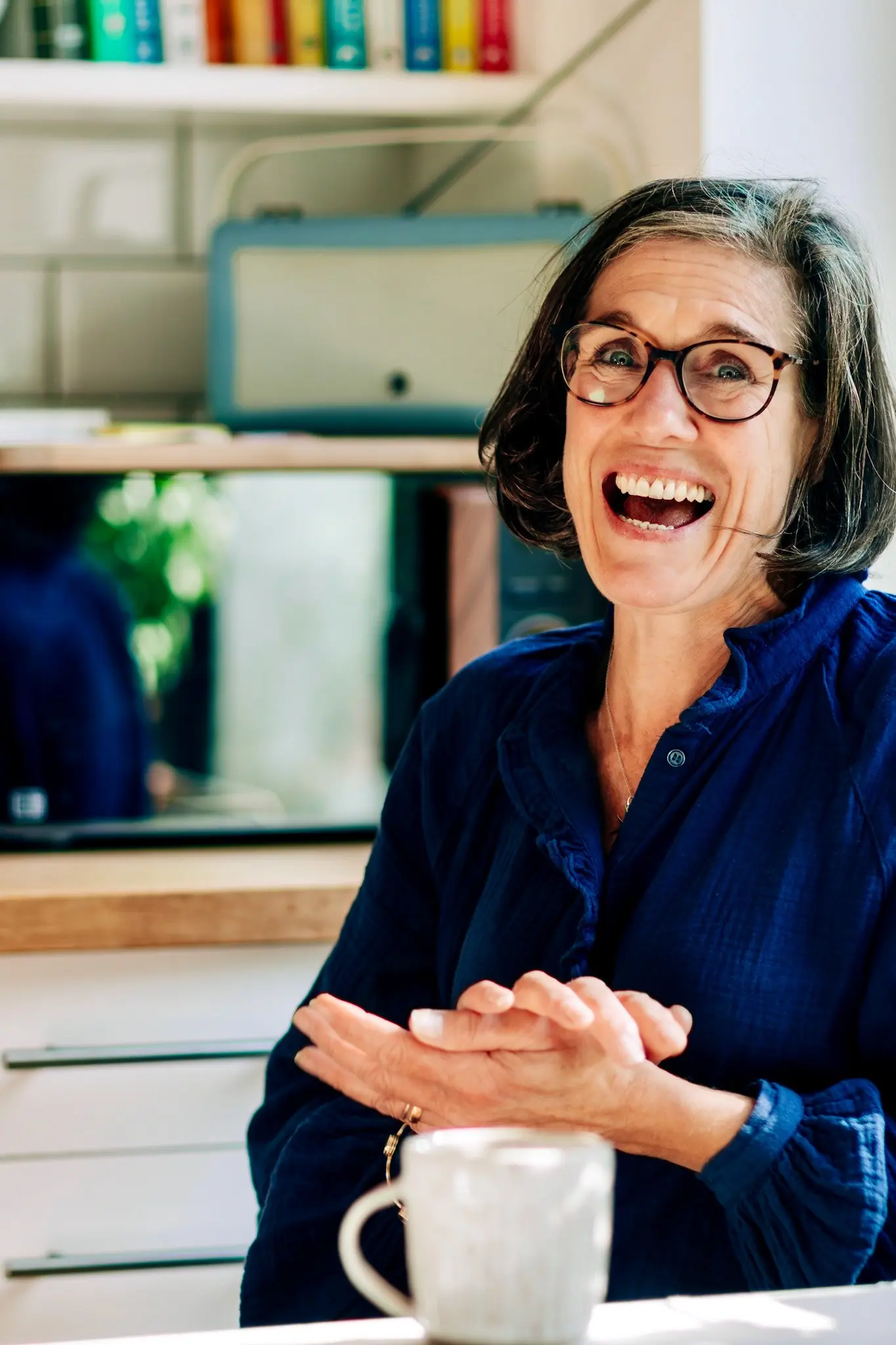 A woman in glasses laughs at a kitchen table, a mug in front of her.