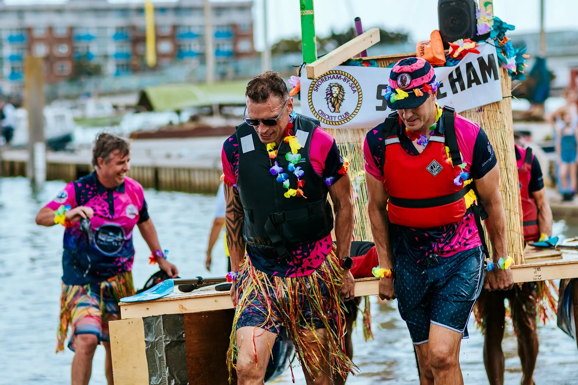 Raft racers in grass skirts and leis carry their homemade raft along the Shoreham riverside.